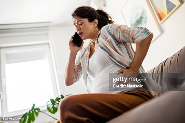 mulher jovem grávida fazendo caretas com as mãos no estômago enquanto usa o telefone inteligente - muscular contraction - fotografias e filmes do acervo