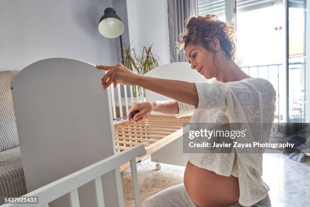 pregnant latin woman assembling a crib in her living room - casa real española fotografías e imágenes de stock