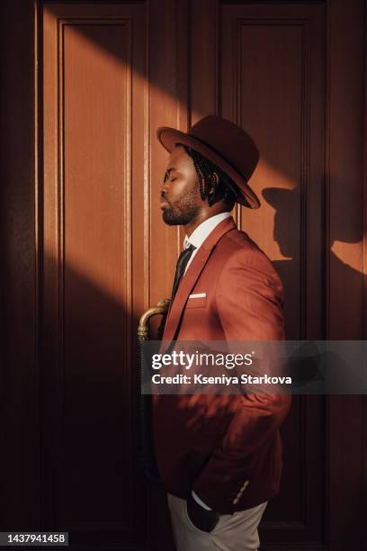 a black young man in a hat with dreadlocks and a brown suit stands against the background of brown doors and looks away, in a ray of sunlight - brown hat imagens e fotografias de stock