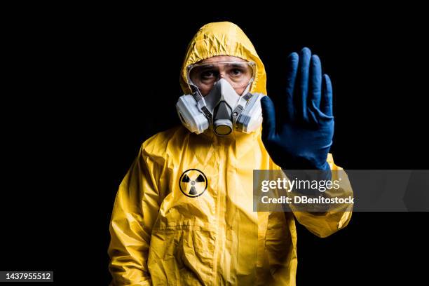 portrait of technician with yellow nuclear protection suit, mask and protective glasses, with the nuclear symbol, making the stop sign, on a black background. concept of nuclear energy and pandemics. - schutzanzug stock-fotos und bilder