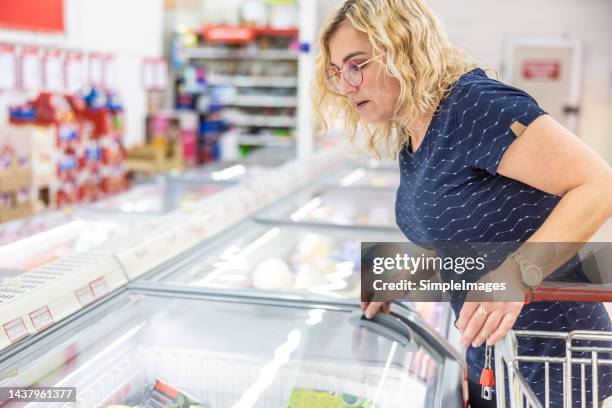 a woman in a grocery store is standing by a freezer and searching for frozen vegetables. - frozen meat stock pictures, royalty-free photos & images