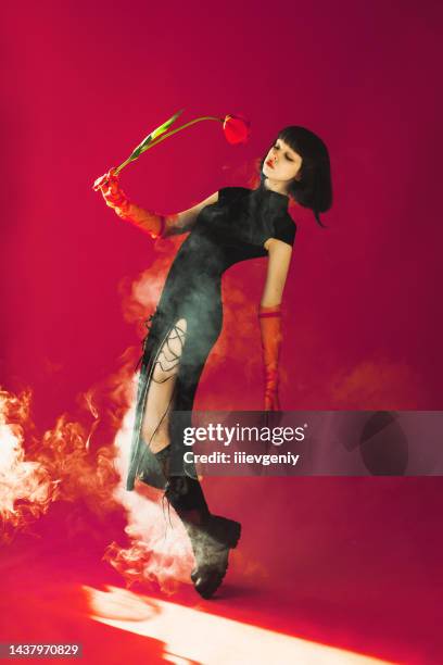 brunette asian woman with tulip in hand posing with smoke in studio on red background. fashion model with pale skin and brown hair in black dress. gloves on hands. style - asian model stock pictures, royalty-free photos & images