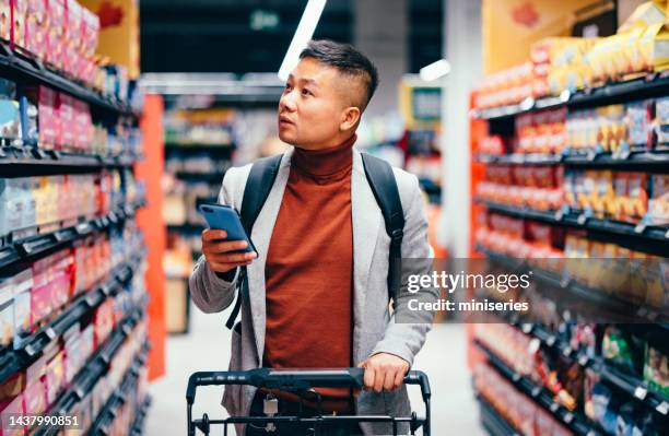 beau mâle asiatique à la recherche de produits d’épicerie dans la liste sur son téléphone portable - consumers retail photos et images de collection