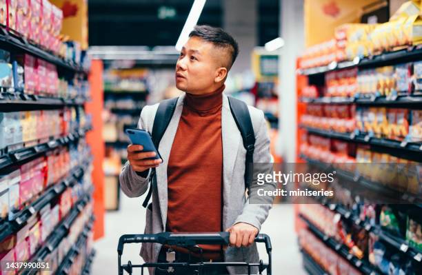 handsome asian male searching for groceries from the list on his mobile phone - supermarkt stock pictures, royalty-free photos & images