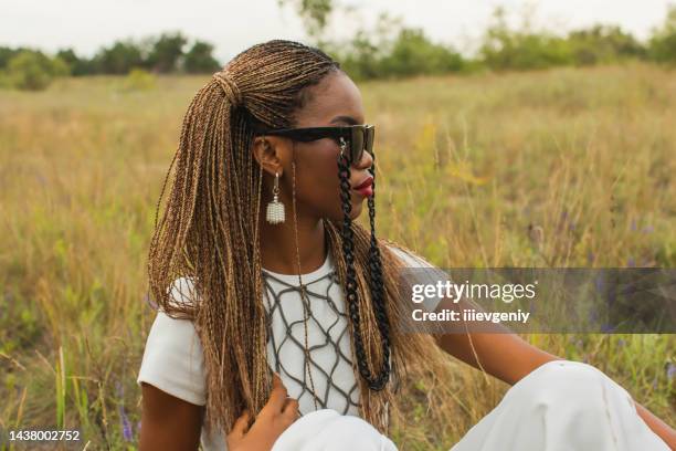 beautiful african woman in black glasses and white shirt and cape on nature background. authentic style. stylish accessories. beauty portrait. earrings. fashion style. woman sit on grass - curly red hair glasses stock pictures, royalty-free photos & images