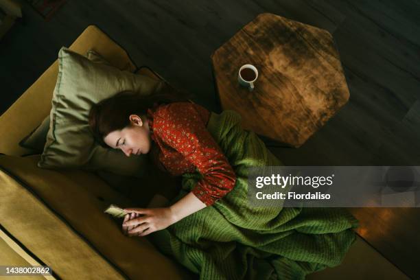 portrait of a red-haired woman in red dress covered with a green blanket lying down on the sofa with cup of tea - romance cover stock pictures, royalty-free photos & images