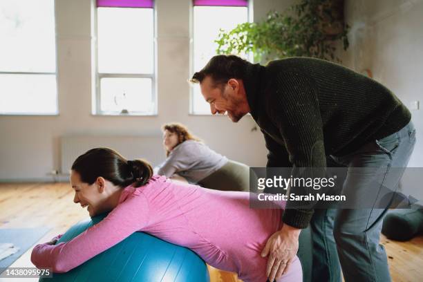 woman in prenatal yoga class resting on birthing ball while partner massages hips - pregnancy class stock-fotos und bilder