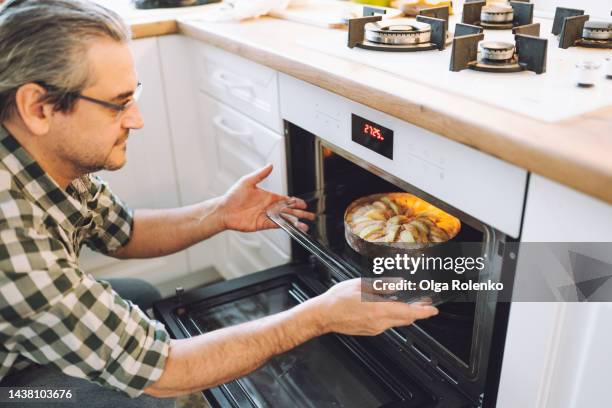 mature househusband pulling out dessert pie from oven. mature man prepare sweet pie or cake at home - house husband stock pictures, royalty-free photos & images