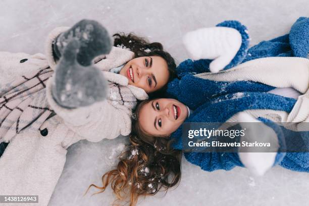 two young women friends in warm clothes are walking, having fun and skating on ice among trees in a snowy forest outdoors in winter. a happy smiling couple laughing and fooling around on a country walk in a winter park on christmas day - ice rink overhead stock pictures, royalty-free photos & images