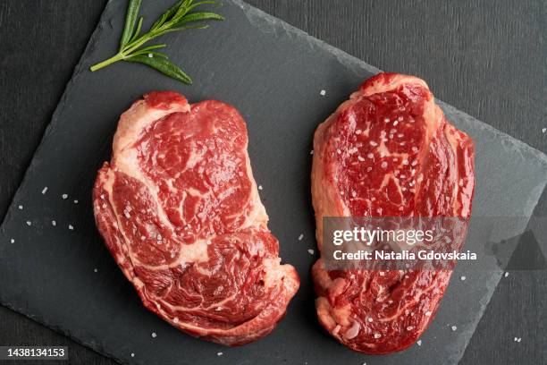 ribeye, rib eye steak, raw beef meat tender marbled striploin steak on dark background, with salt, garlic clove and spices rosemary on black stone board. uncooked sirloin. preparation for cooking - rosbif photos et images de collection