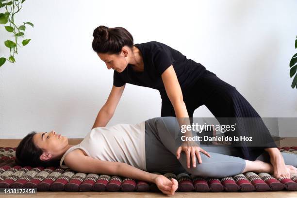 latina masseuse applying thai massage technique to young female client to stretch out hips and spine. assisted spinal twist lying down on futon. - thai massage stock-fotos und bilder