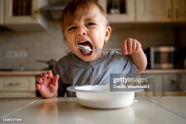 baby boy eating porridge with spoon in kitchen - boy eating cereal foto e immagini stock