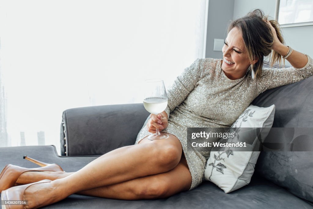 Happy woman in cocktail dress holding wineglass on sofa at home