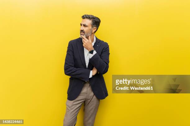 thoughtful businessman with hand on chin standing against yellow wall - veste jaune photos et images de collection