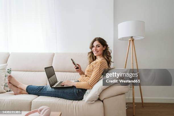 smiling young woman with laptop and mobile phone on sofa - businesswoman barefoot stock-fotos und bilder