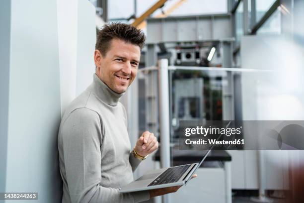 smiling businessman holding laptop leaning on wall in industry - schweiz business stock-fotos und bilder