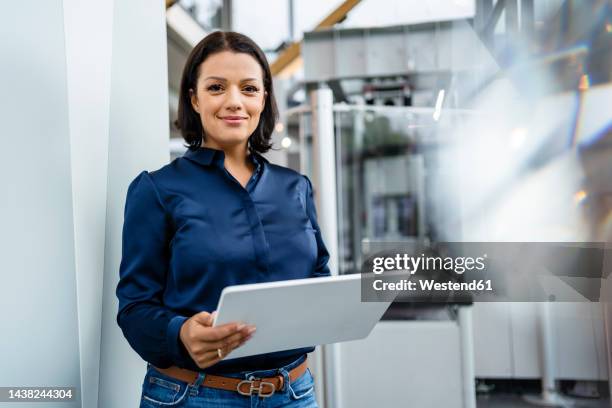 smiling businesswoman holding tablet pc standing by wall in industry - industry 40 stock pictures, royalty-free photos & images