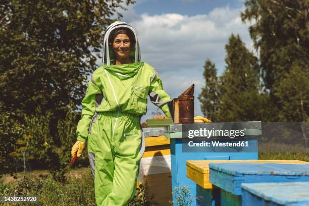 smiling beekeeper with bee smoker standing by wooden boxes on sunny day - apiculture stock pictures, royalty-free photos & images