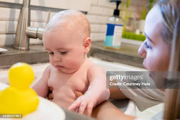 young adult mother is giving her infant son a bath in the kitchen sink at home and wrapping him in a towel - baby bath toys stock pictures, royalty-free photos & images