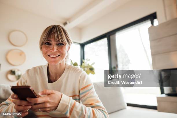 happy woman holding mobile phone at home - in den vierzigern stock-fotos und bilder