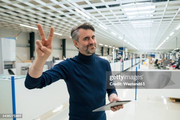smiling businessman with tablet pc showing peace gesture in industry - gestikulieren mit den händen stock-fotos und bilder