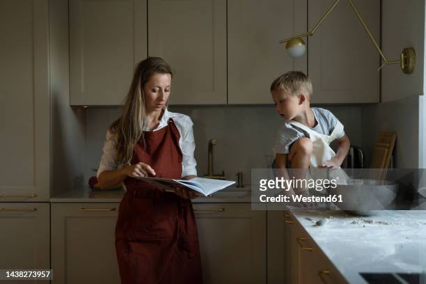 mother reading recipe book with son sitting on kitchen counter - cookbook stock pictures, royalty-free photos & images