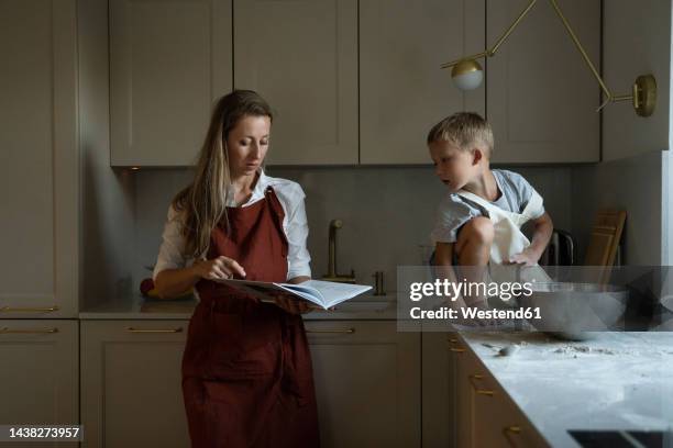 mother reading recipe book with son sitting on kitchen counter - avental imagens e fotografias de stock