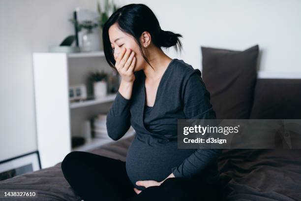 young asian pregnant woman sitting on her bed, feeling nausea and covering her mouth, suffering from morning sickness. pregnancy health, wellness and wellbeing concept - morning sickness stock-fotos und bilder