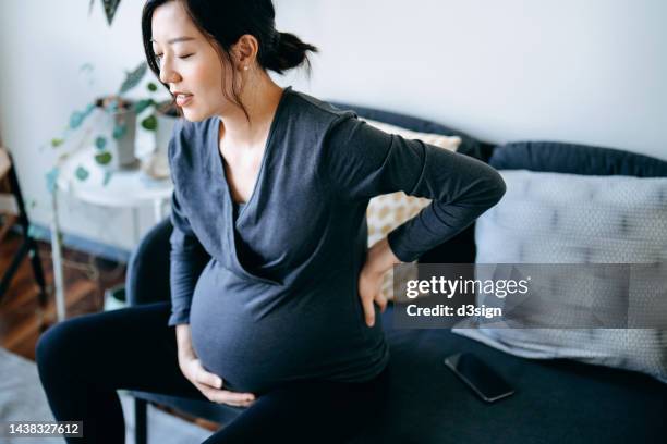 young asian pregnant woman touching her baby bump and lower back, suffering from backache. pregnancy health, wellness and wellbeing concept - morning sickness stockfoto's en -beelden