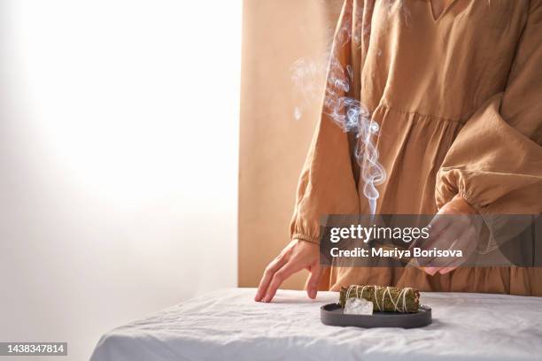 the hands of a girl in a beige dress hold a smoking palo santo stick against the background of ritual things: a rock crystal stone and a twist for fumigation. - smudging ceremony stock pictures, royalty-free photos & images