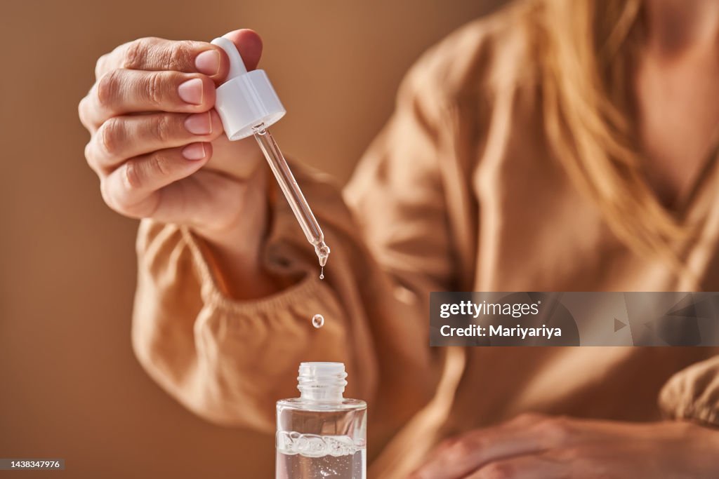 A girl in a beige dress holds a pipette with a drop of serum in her hands.