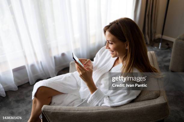 junge frau lächelt, während sie ihr handy überprüft und im sessel genießt - checking in at hotel stock-fotos und bilder