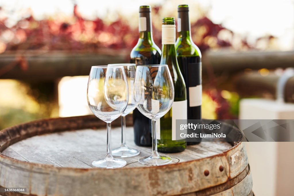 Empty wine glasses, wine and barrel in a vineyard for wine tasting, celebration or party in nature. Winery, bottles and luxury alcohol on a wooden drum for dinner, special banquet or event on a farm.