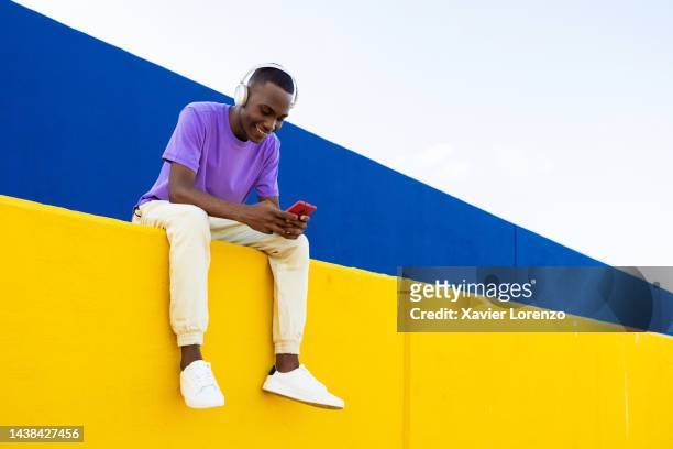 cheerful young colombian hispanic latin man with headphones having fun using mobile phone while relaxing outdoors sitting on yellow wall - social media, technology and millennial people concept - young man listening to music on smart phone outdoors stockfoto's en -beelden