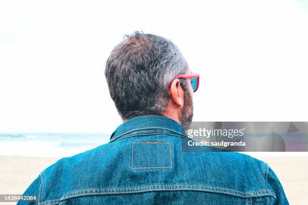 portrait of gray-haired man with denim jacket looking at the sea, rear view - man rear view grey hair closeup stock pictures, royalty-free photos & images