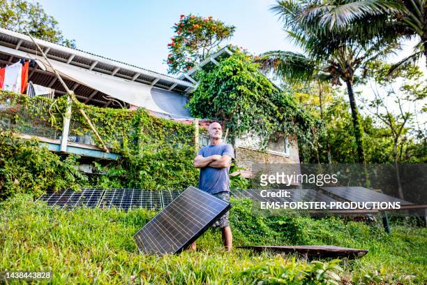 active, male senior is standing beside his solar panels in his jungle garden - 自给自足 個照片及圖片檔