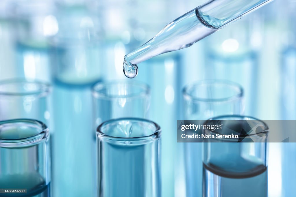Laboratory glassware with dropper dripping liquid into test tube with light blue liquid close up macro photography.