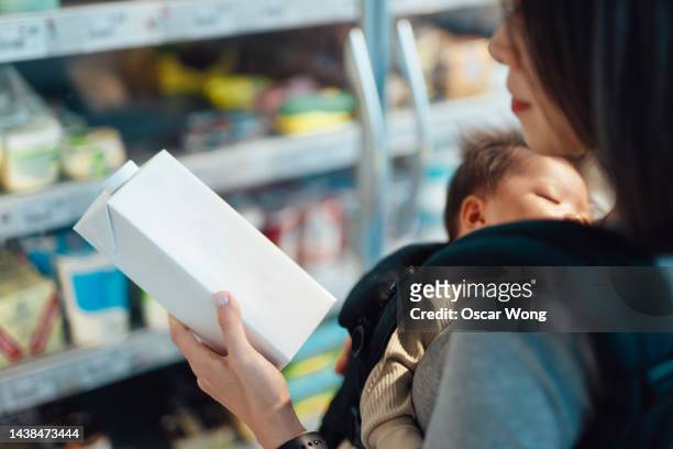 over the shoulder view of young woman shopping plant-based milk at supermarket while holding her daughter in baby carrier - mom buying milk photos et images de collection