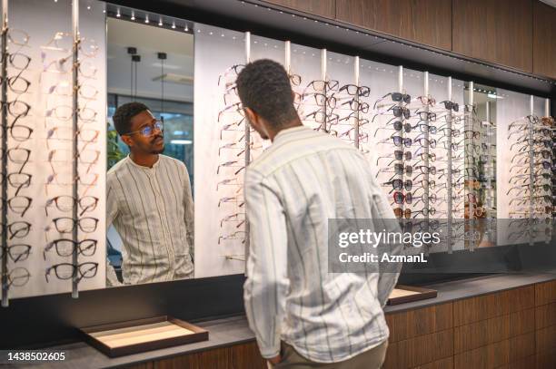 attractive mid adult black male looking at eyeglasses in the mirror - choosing eyeglasses stock pictures, royalty-free photos & images