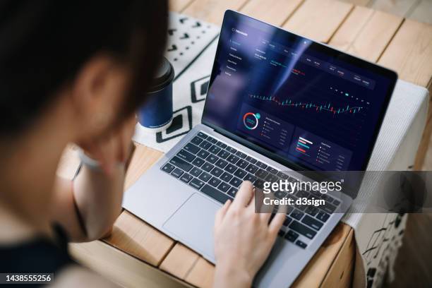 over the shoulder view of young asian woman managing finance and investment, analyzing stock market data on laptop at desk. stock exchange, banking, finance, investment, financial trading concept. smart banking with technology - hot desking arbeitsplatz stock-fotos und bilder