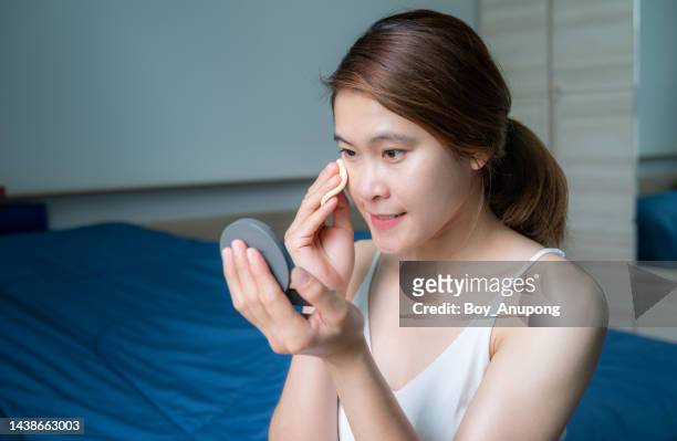 portrait of asian woman while applying foundation power puff on her face to cover flaws and change the natural skintone. - powder puff stock pictures, royalty-free photos & images