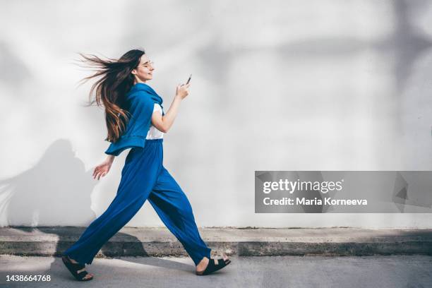 woman using mobile phone while walking in front of concrete wall. - peatón fotografías e imágenes de stock