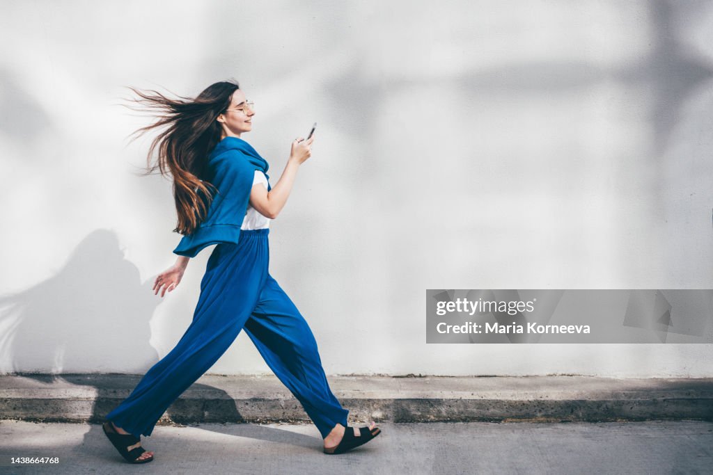 Woman using mobile phone while walking in front of concrete wall.