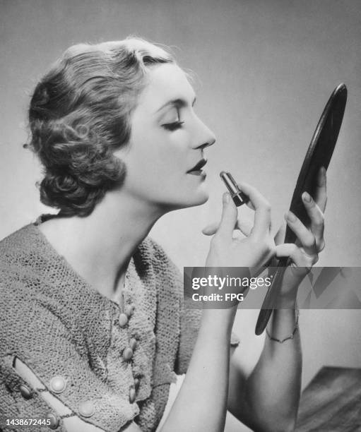 Woman holding a handheld mirror with her left hand, applying 'Rouge Mandarine' lipstick with her right hand, United States, circa 1935.