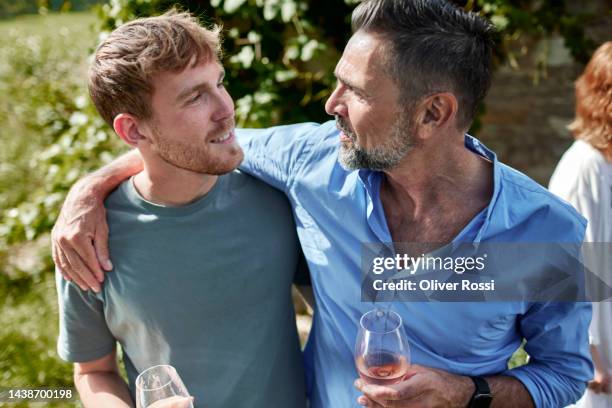 mature man with wine glass embracing young man in garden - reencuentro fotografías e imágenes de stock