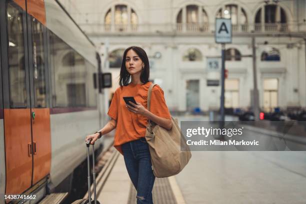 una joven y bella mujer española está usando un teléfono móvil en una estación de tren mientras espera para entrar en un vagón de tren - estación de tren fotografías e imágenes de stock