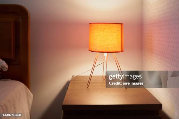 table lamp lit on a small table in the corner of a room with a white wall next to a bed - lámpara eléctrica fotografías e imágenes de stock