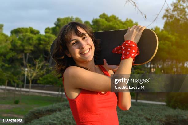 professional skateboarder girl in camisole carrying a skateboard on her left shoulder at in the park. lifestyle concept of a young caucasian girl in a red camisole carrying her skateboard on her shoulder in a green park - cami parker bildbanksfoton och bilder