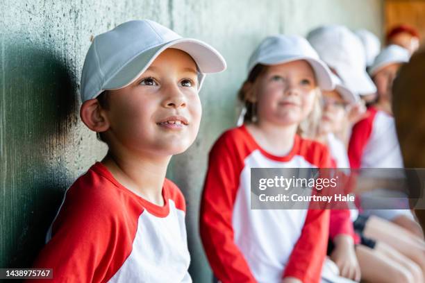 cute little boy smiles while sitting in dugout with little league team during game - girl baseball cap stock pictures, royalty-free photos & images