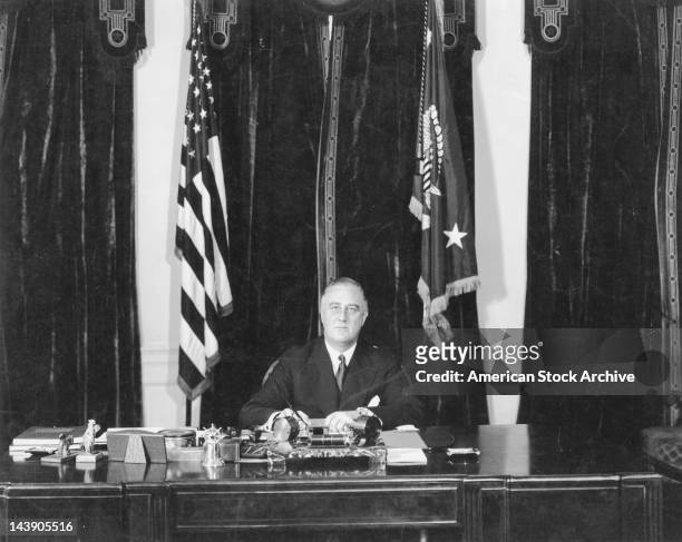American President Franklin Delano Roosevelt seated at his desk in front of American flags, circa 1940.